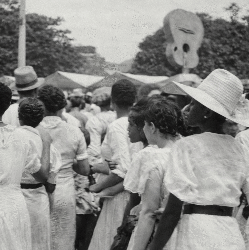 A fotografia retrata uma cena urbana antiga da tradicional Lavagem do Bonfim. Em primeiro plano, há um grande grupo de homens reunidos ao redor de uma carroça. As pessoas na multidão ao redor do veículo usam roupas claras, ternos leves, camisas, calças sociais e chapéus. Ao redor, outras pessoas observam a cena, e também há crianças próximas ao grupo.